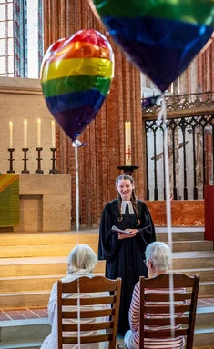 Zwei Frauen sitzen auf Holzstühlen mit dem Rücken zur Kamera, vor Ihnen steht eine Frau mit schwarzem Talar und weißer Schleife. 