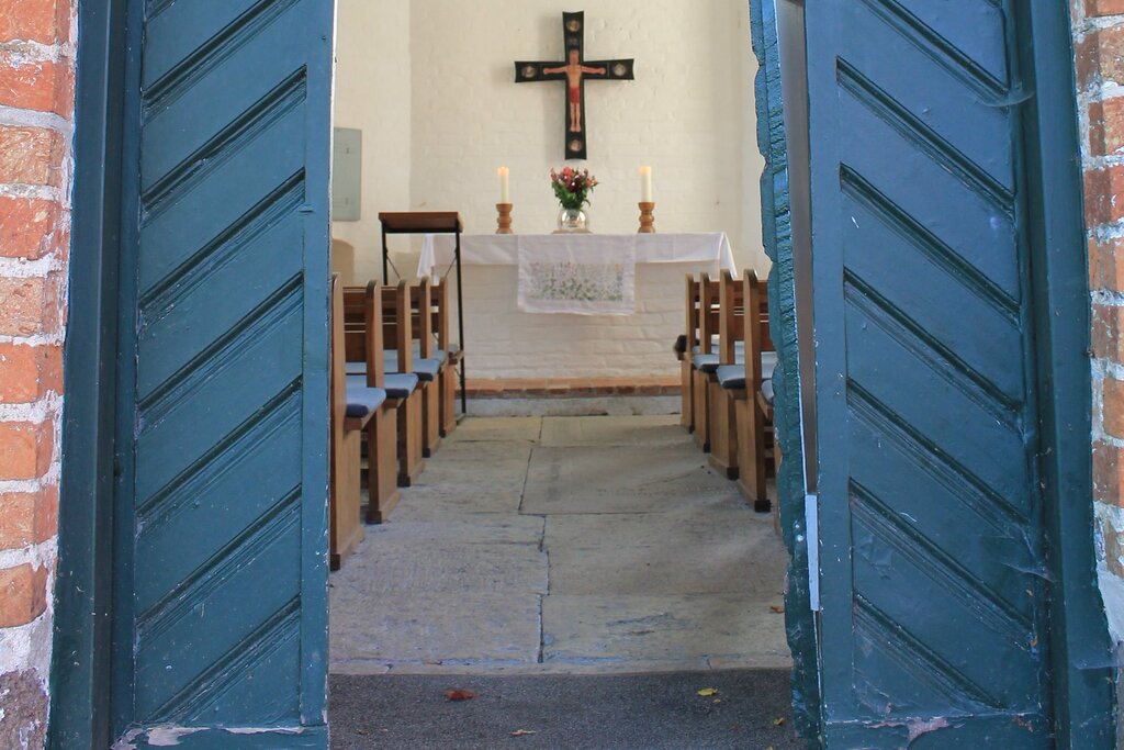 Eine offene blaue Kirchentür gibt den Blick frei zu einem Kirchenaltar auf dem zwei große, weiße, brennende Kerzen zu sehen sind. Zwischen den Kerzen steht eine runde Glasvase mit Blumen, darüber hängt ein Altarkreuz. Im vordergrund sind braune Holz-Kirchenbänke mit blauen Sitzauflagen zu sehen.