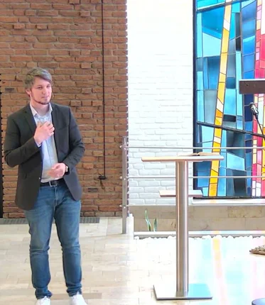 Ein Mann steht im Vordergrund neben einem Stehpult, dahinter ein dunkles Holzkreuz und ein buntes Glasfenster