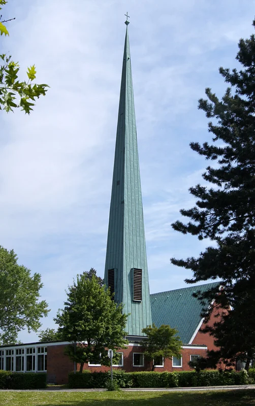sehr hoher, spitzer Glockenturm mit Kupferblech, rechts Kirche, unten Gemeinderäume          