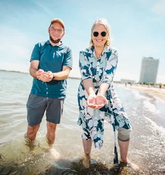 Zwei Personen stehen in der Ostsee, haben Wasser mit den Händen geschöpft; im Hintergrund ist das Travemünde Maritim zu sehen.