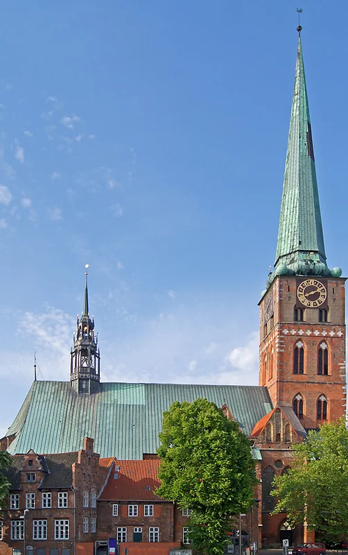 St.-Jakobi-Kirche Lübeck vom Klingenberg fotografiert. Der Turm ragt in den blauen Himmel. Es ist eine Seitenansicht der Kirche, rechts der Turm, links das Kirchenschiff. Vor der Kirche stehen links zwei Bäume mit grünen Blättern.