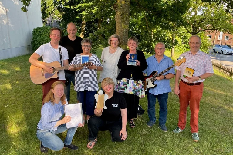 Neun Menschen stehen und sitzen zusammen auf einer Wiese und gucken freundlich in die Kamera.