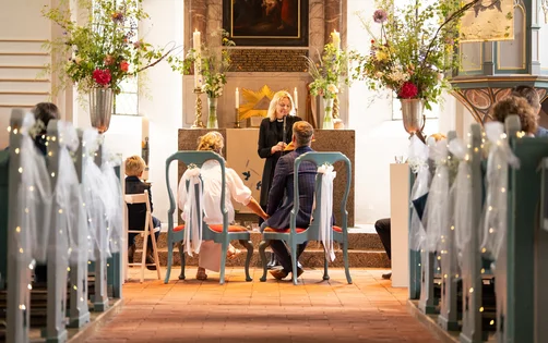 In einer Kirche mit weißen schleiern geschmückte Bänke. Ganz hinten zwei Personen auf Stühlen mit Rücken zum Fotografen. Davor eine Frau in Talar vor einem Altar. Grünpflanzen und Blumen sind an den Wänden zu sehen.