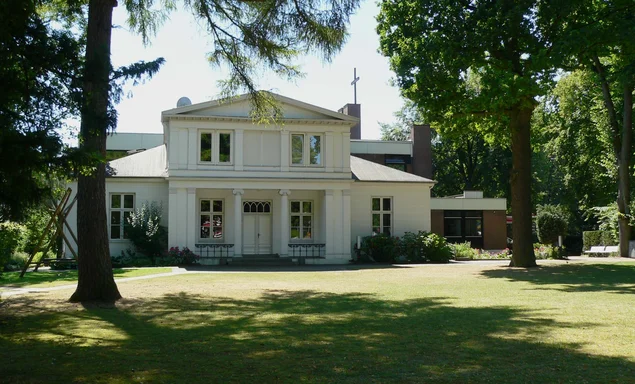Ein weißes Haus mit grüner Wiese im Vordergrund und zwei Bäumen links und rechts
