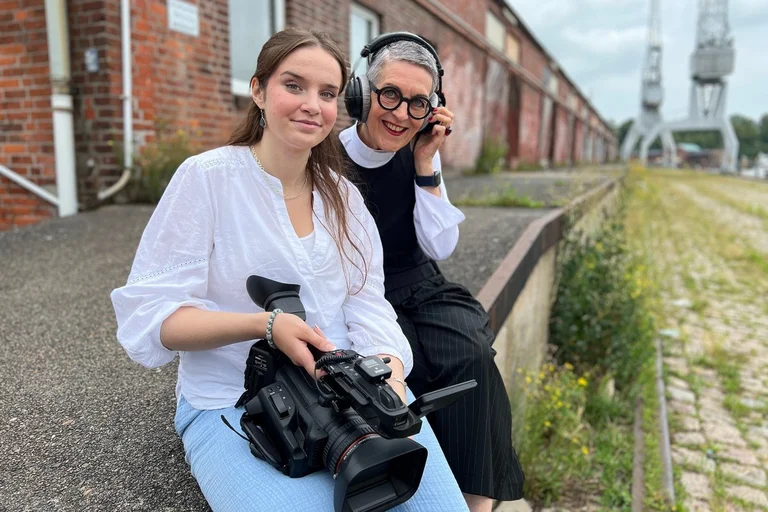 Zwei Frauen: links eine junge Frau mit einer großen Filmkamera auf dem Schoß, rechts eine Frau mit Kophörern