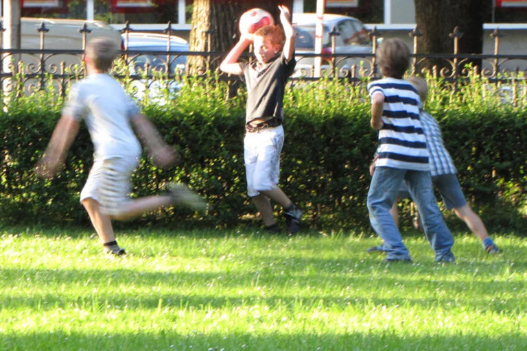 Vier Jungen spielen auf einem Rasen der Teils sonnenbeschienen ist. Der zweite Junge von links hält einen Ball über seinem Kopf