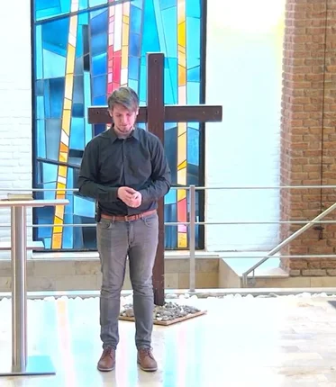 Ein Mann steht betend vor einem Holzkreuz und hat die Augen geschlossen - Neben ihm steht ein Stehtisch mit einer Bibel darauf. hinter ihm ist ein Becken und ein Handlauf zu sehen, ebenso ein buntes Glasfenster