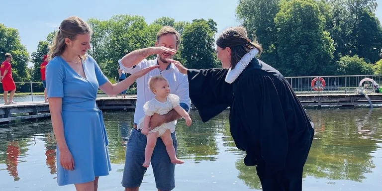 Eine Pastorin tauft ein kleines Mädchen im Krähenteich. Der Vater hält es hoch, die Mutter schaut zu freut sich. Beide Eltern halten ihre Hände als Segen über den Kopf des Kindes. 