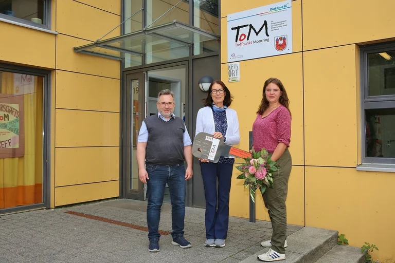 Ein Mann und zwei Frauen stehen vor einem gelben Gebäude und halten einen großen Schlüssel aus Pappe.