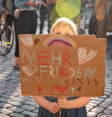 Kind hinter einem Schild mit der Aufschrift "Mehr Frieden, weniger Nazis"