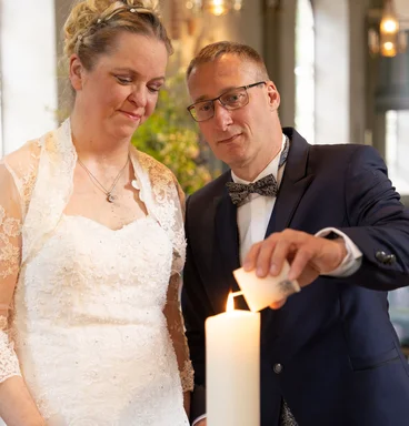 Ein Brautpaar Mann und Frau in Festkleidung. Der Mann zündet mit einer kleinen weißen Kerze eine große an.