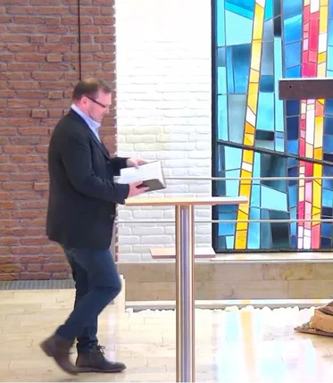Ein Mann geht zu einem Stehpult, er hält ein offenes Buch in den Händen, rechts steht ein großes Holzkreuz, im Hintergrund ist ein buntes Kirchenfenster zu sehen
