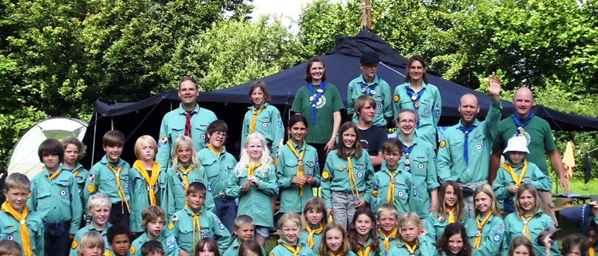eine groß Gruppe Pfadfinder steht und sitzt in vier Reihen und schaut in die Kamera; im Hintergrund sind grüne Bäume und ein Gebäude zu sehen