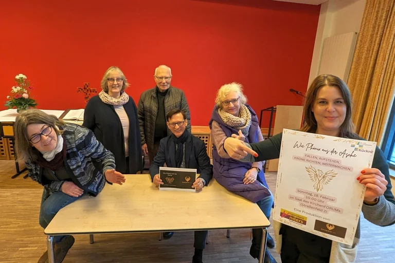 Sechs Frauen und Männer stehen um einen Tisch und schauen in die Kamera.