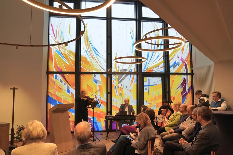 Ein Raum mit großen bunten Fenstern, vielen sitzenden Menschen und drei musizierendenMusikern.