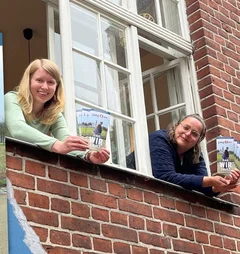 Zwei Frauen schauen aus einem Fenster und halten Flyer in den Händen.