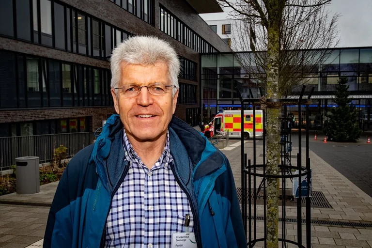 Ein Pastor steht vor dem Portal eines Krankenhauses und lächelt in die Kamera. 