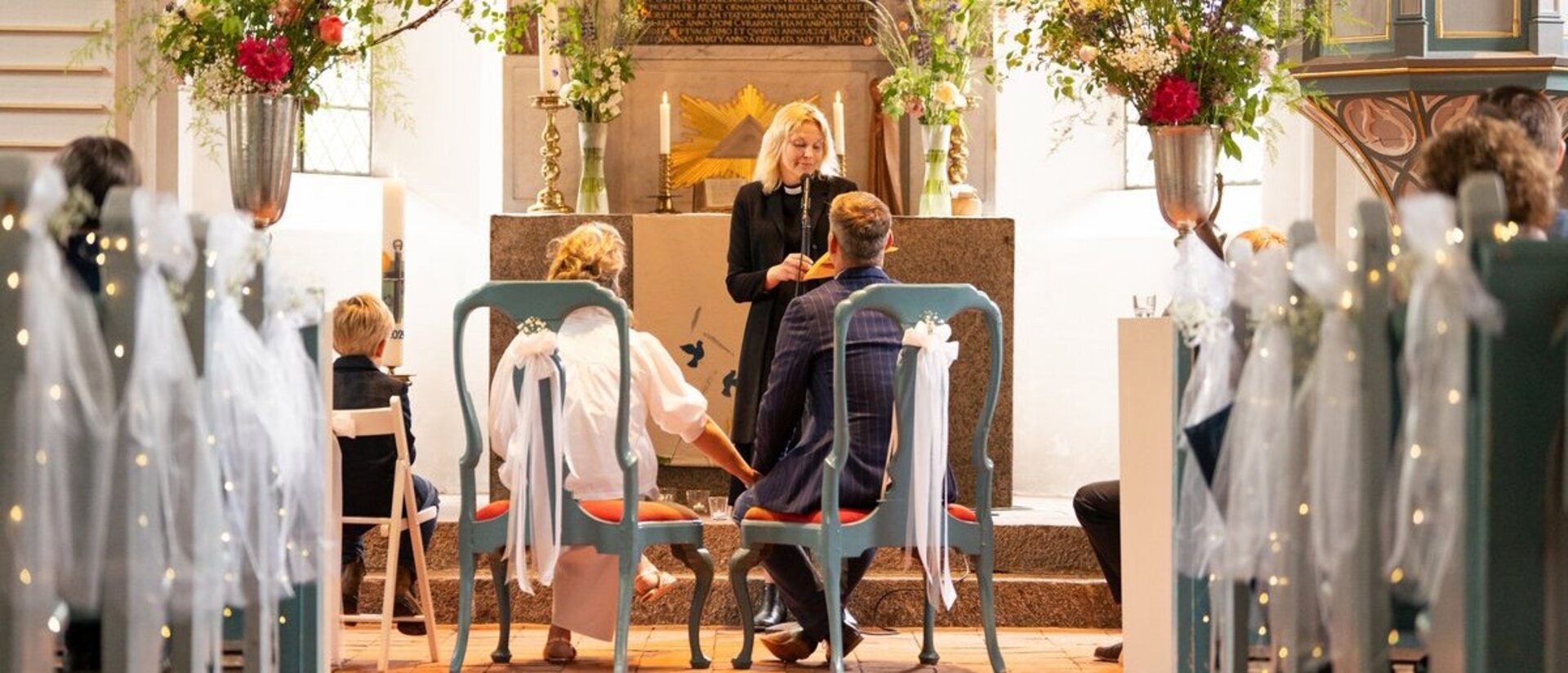 Blick in den geschmückten Mittelgang einer Kirche mit Brautpaar und Pastorin im Altarraum..