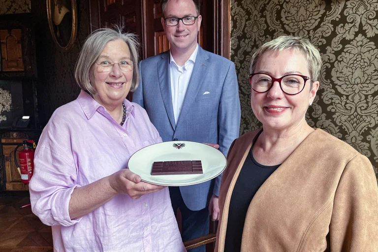 Zwei Frauen und ein Mann stehen in einem Raum und zeigen eine Tafel Schokolade