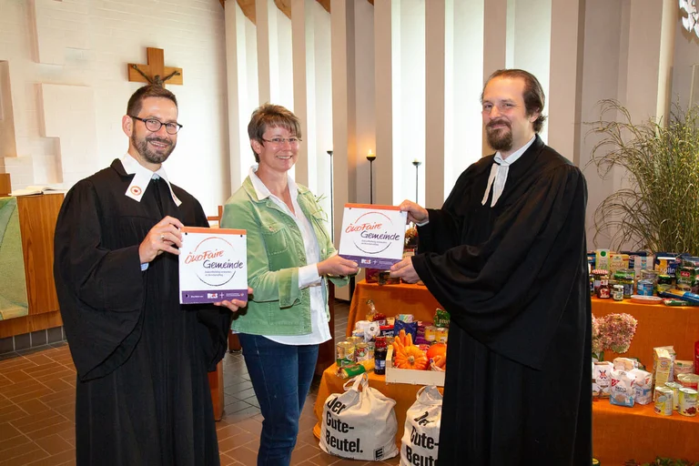 Drei Personen stehen mit einem Schild lächelnd in einer Kirche.