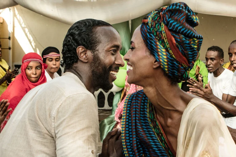 Ein Mann und eine Frau sitzen sich lächelnd gegenüber, ringsum sind weitere fröhliche Menschen zu sehen. 