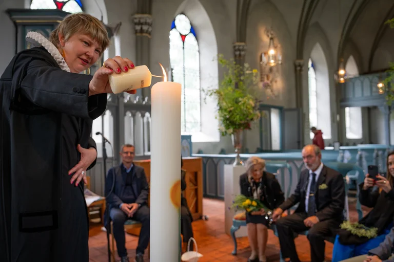 Eine Pastorin zündet eine große weiße Kerze an und davor sitzen Traupaare auf Stühlen.