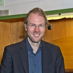Junger Mann (Organist) sitzt mit dem Rücken zu einer Orgel und lächelt in die Kamera