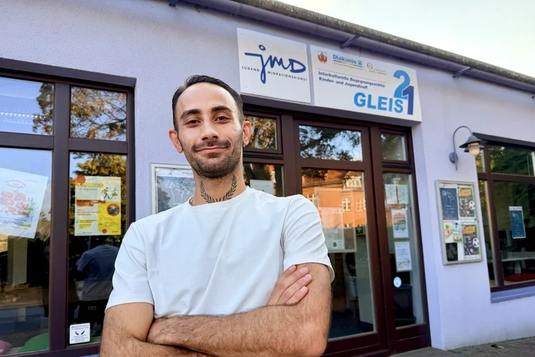 Ein Mann mit weißem T-Shirt und dunklen kurzen Haaren und Drei-Tage-Bart steht vor einem Gebäude mit großen Türen und Fenstern. Er hat die Arme vor sich verschränkt.
