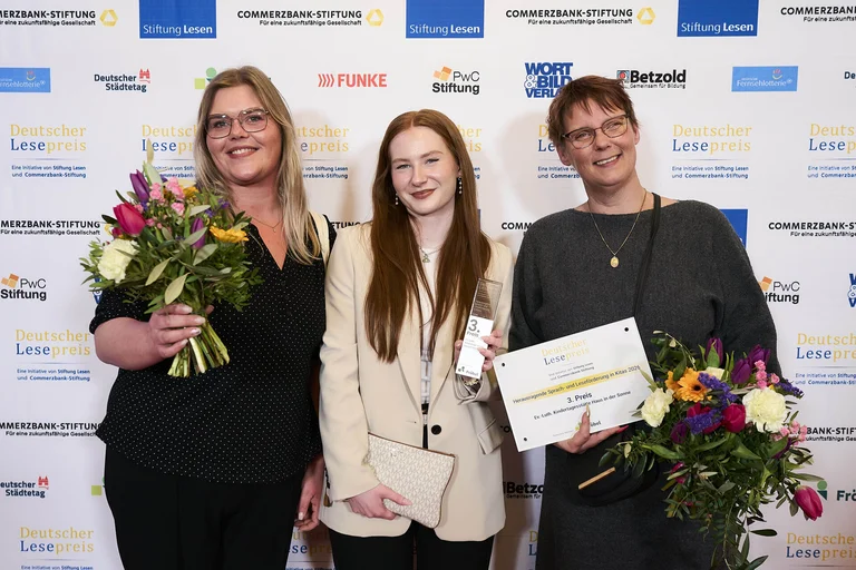 Drei Frauen stehen vor einer Wand und halten Blumen und eine Urkunde.