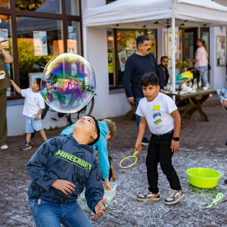 Mehrere Kinder und Erwachsene mit Migrationshintergrund. Ein Kind dukt sich unter einer großen Seifenblase