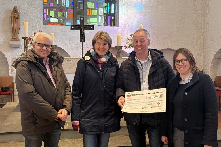 Zwei Frauen und zwei Männer stehen in einer Kirche und halten einen großen Scheck in die Kamera.