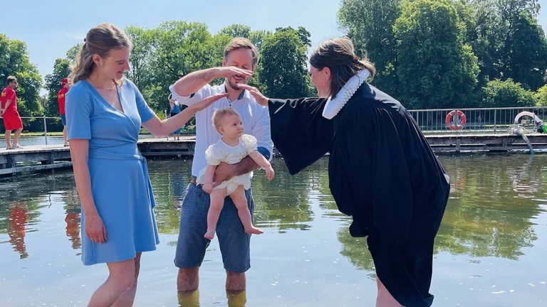 Eine Pastorin tauft ein kleines Mädchen im Krähenteich. Der Vater hält es hoch, die Mutter schaut zu freut sich. Beide Eltern halten ihre Hände als Segen über den Kopf des Kindes. 