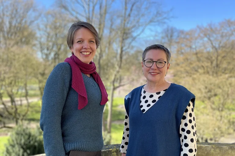 Zwei Frauen stehen im Freien und lächeln in die Kamera.