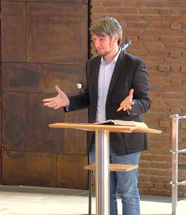 Ein Mann steht vor einem Pult und hält die Arme vor sich geöffnet und schaut nach vorne