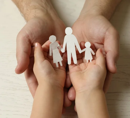 Ein paar offene Kinderhände alten eine Familie aus Mutter-Vater-Kind-Scherenschnittfamilie (weißes Papier) und Erwachsenenhände halten die Kinderhände darunter