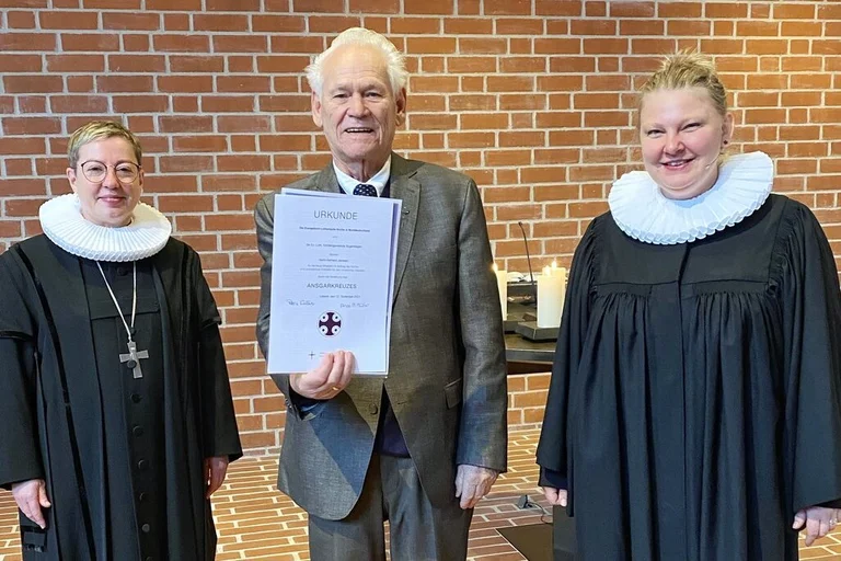 Drei Personen stehen in einer Kirche und lächeln in die Kamera. In der Mitte steht ein Mann mit einer Urkunde.