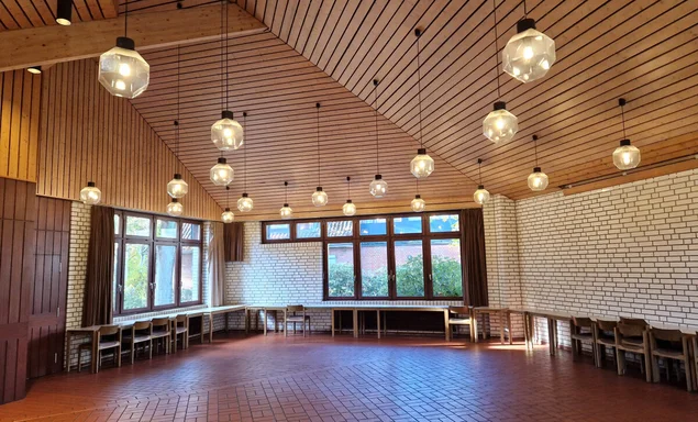 Blick in den großzügigen Gemeindesaal im Standort Düneberg mit Holzvertäfelter Decke und von der Decke herabhängenden Kugelleuchten. Die Fensterfronten an den beiden außenänden lassen viel licht hinein, der Boden ist gefließt. 