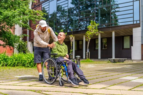 Ein Mann mit Kappe hält die Griffe eines Rollstuhles und lächelt auf den Mann im Rollstuhl. Dieser lächelt vergnügt, er hat eine Behinderung.