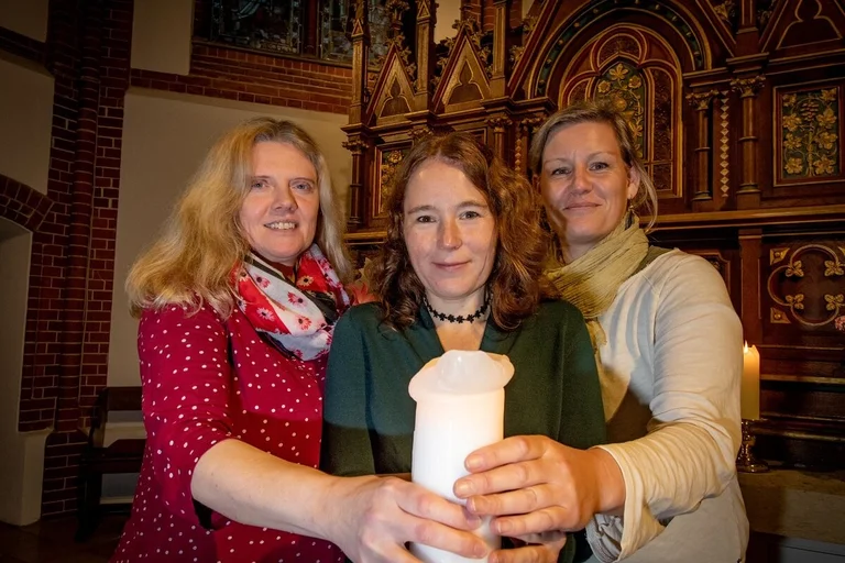 Drei Frauen stehen in einer Kirche und halten gemeinsam eine Kerze. 