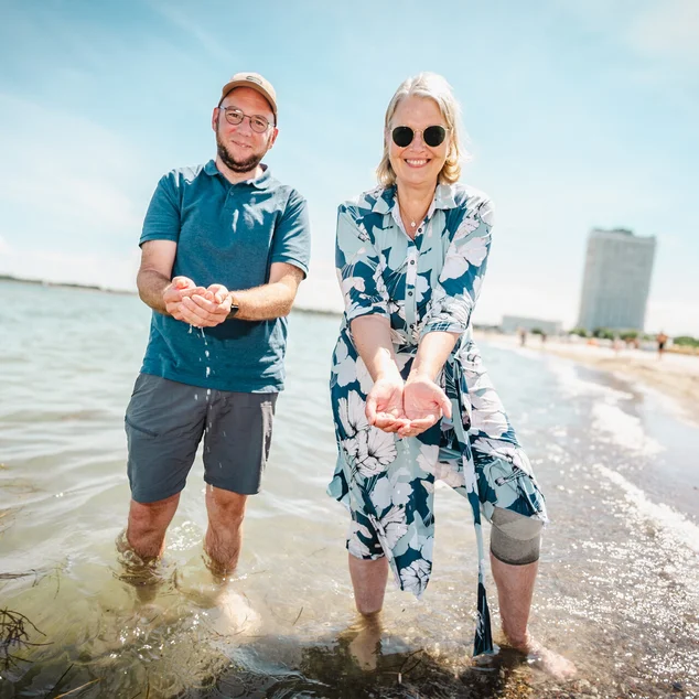 Zwei Personen stehen in der Ostsee, haben Wasser mit den Händen geschöpft; im Hintergrund ist das Travemünde Maritim zu sehen.