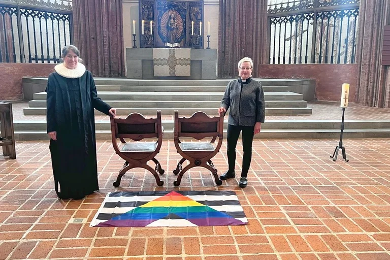 Zwei Personen stehen in einer Kirche neben zwei freien Stühlen. 