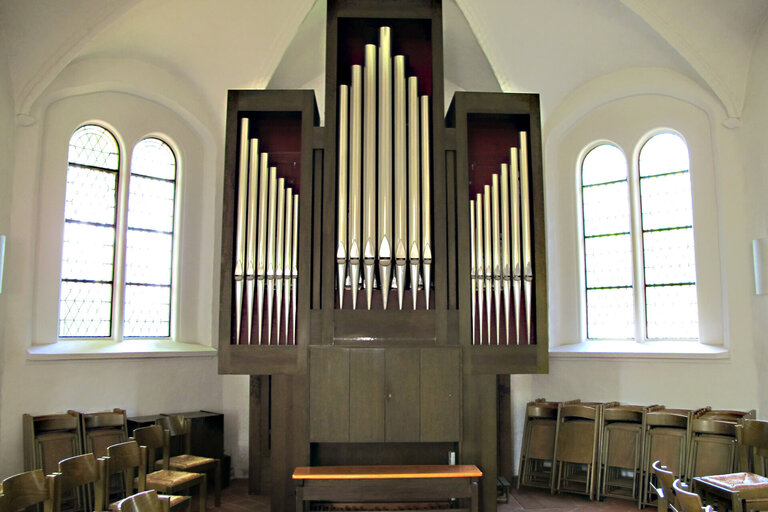 Eine Orgel mit drei Orgelpfeifenkästen und einer Sitzbank steht vor den Kirchenfenstern in einer Kirche, links und rechts stehen Klappstühle, teils zugeklappt an den Wänden gelehnt, teils offen nebeneinander stehend.