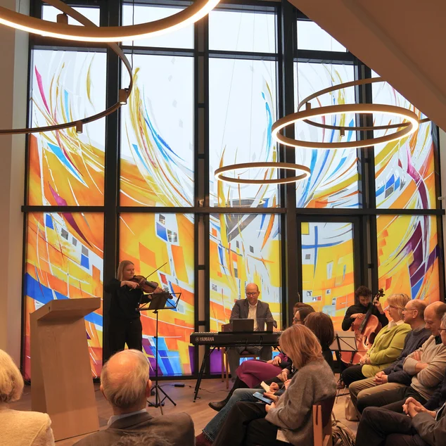 Ein Raum mit großen bunten Fenstern, vielen sitzenden Menschen und drei musizierendenMusikern.
