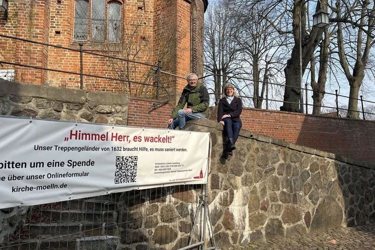 Alte Kirchenmauer mit einem Spendenbanner und zwei Personen, die auf der Mauer sitzen.