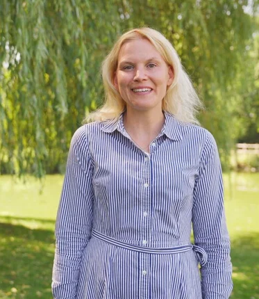 Eine blonde Frau mit gestreiften Kleid steht vor einem Baum.