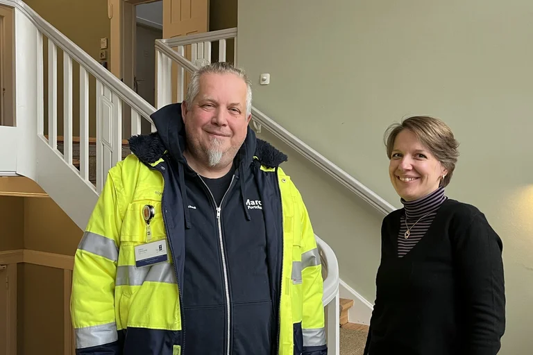 Ein Mann mit gelber Warnjacke steht neben einer Frau mit schwarzem Pullover vor einem weißen Treppengeländer.