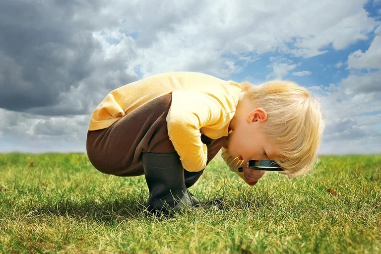 Ein kleiner Junge erkundet die Natur draußen, indem er Gras durch eine Lupe betrachtet. Er ist in der Hocke und trägt einen gelben Pullover, eine braune Hose und grüne Gummistiefel.