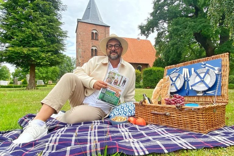 Ein mann sitzt auf einer Picknickdecke vor einer Kirche und schaut in die Kamera. 
