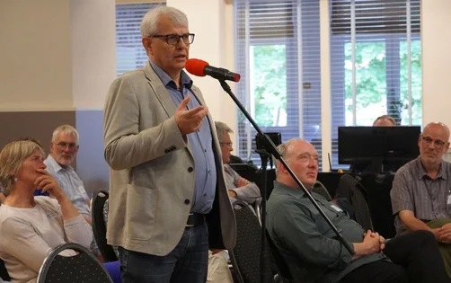 Ein Mann mit grauen Haaren steht an einem Mikrofon und spricht.